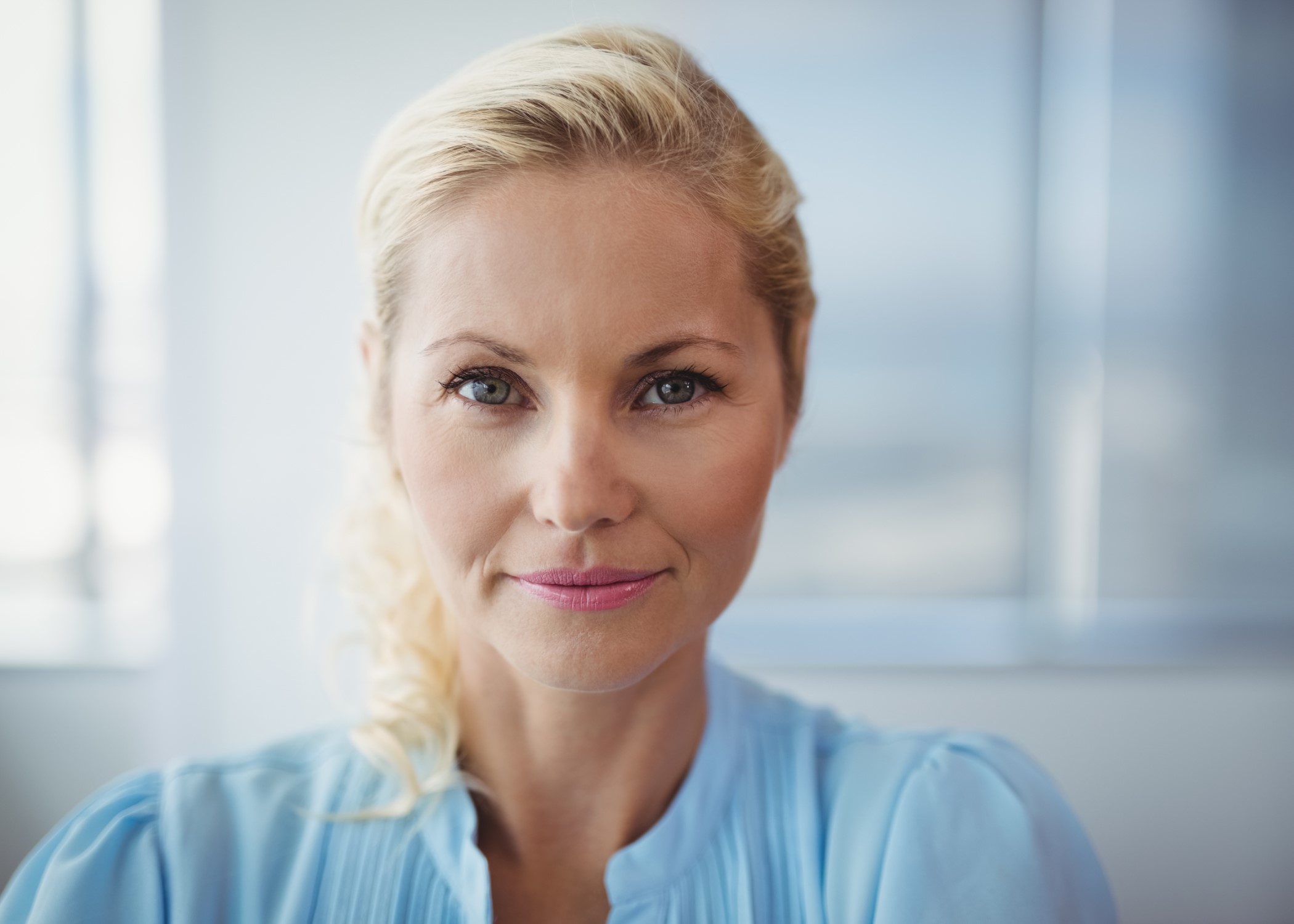 Close-up portrait of a smiling blonde woman with light blue eyes, wearing a light blue blouse. She exudes confidence and professionalism in a well-lit office setting.