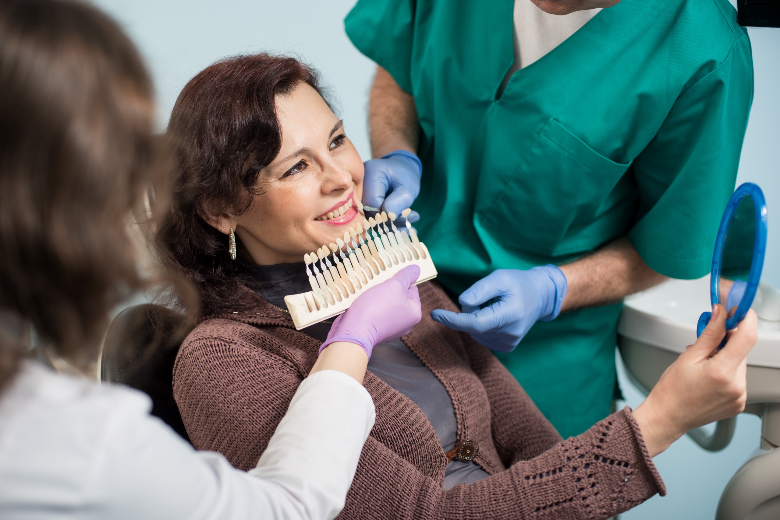 A dentist and assistant helping a female patient choose the right tooth color for dental veneers using a color shade guide in a dental office.