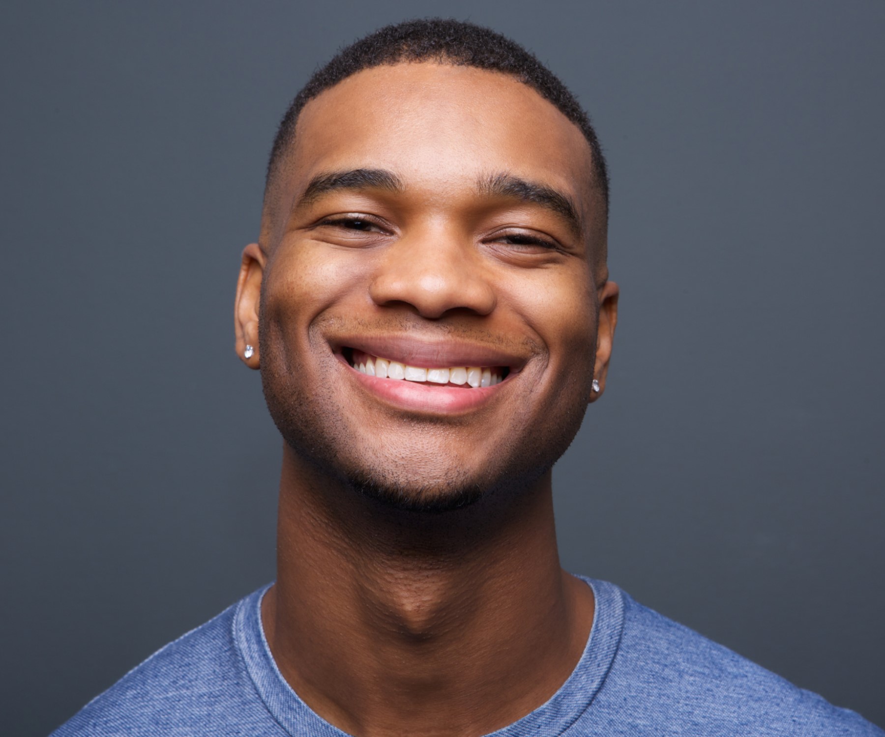 Close-up of a happy black man smiling confidently in front of a gray background, showcasing positivity and warmth.