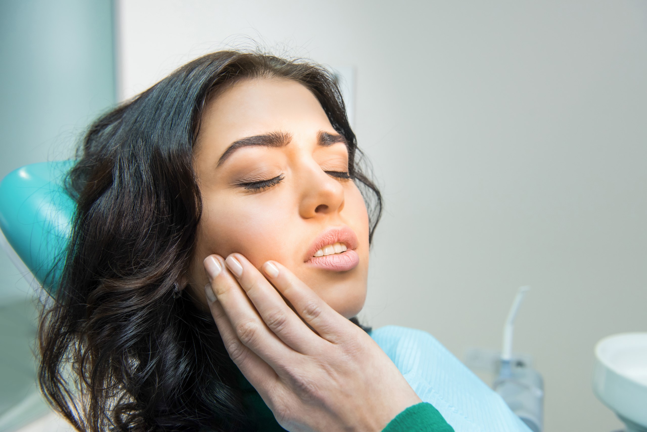 Young woman in a dental chair experiencing TMJ symptoms, holding her cheek in discomfort during a dental appointment.